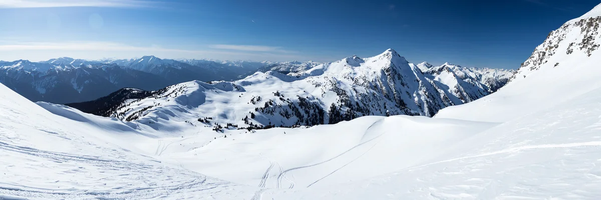 Panorama of the Foggy Day riding area, sled tracks across an open snow bowl with the Selkirks beyond